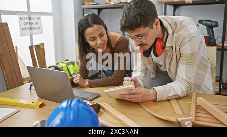 Un uomo e una donna collaborano in un'officina di falegnameria, facendo riferimento a un notebook e annotando su un blocco note tra gli utensili in legno. Foto Stock