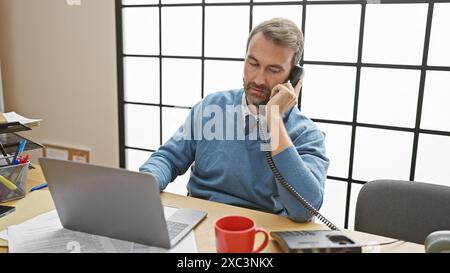 Uomo ispanico di mezza età con barba in camicia con bottoni e maglione che parla al telefono mentre lavora al computer portatile in ufficio. Foto Stock
