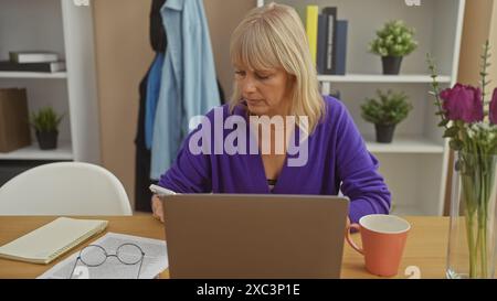 Una donna concentrata di mezza età sfoglia il suo telefono a casa con un laptop, occhiali e caffè sul tavolo. Foto Stock