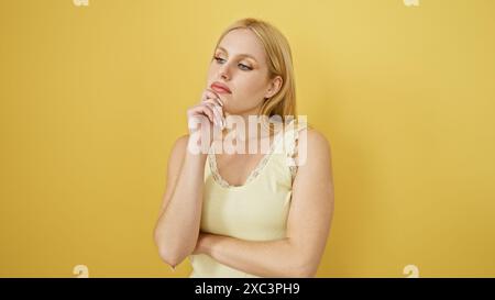 Una giovane donna bionda con un top senza maniche che posa contro un muro giallo, trasuda bellezza e contemplazione casuali. Foto Stock