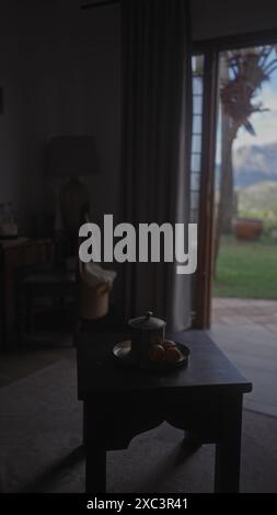 Un ambiente interno con un'atmosfera tranquilla, caratterizzato da un tavolo pieno di frutta, pentola di ceramica, tenda, sedia e vista su un giardino esterno. Foto Stock