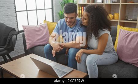 Un uomo e una donna che si accoccolano su un divano in un accogliente soggiorno con laptop e cuscini colorati. Foto Stock