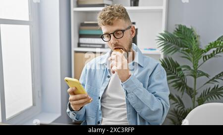 L'uomo barbuto in occhiali mangia un panino mentre controlla il suo smartphone in un ufficio moderno. Foto Stock