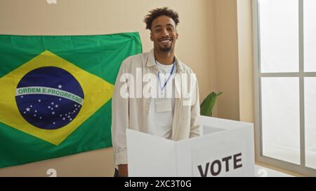 Un giovane afro-americano con la barba si trova in una sala di votazione, posato accanto a una urna con una bandiera brasiliana sullo sfondo. Foto Stock