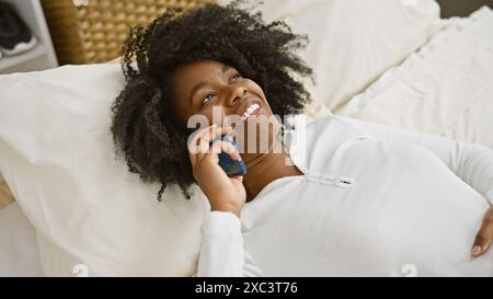 Donna afroamericana sorridente che si rilassa su un letto e fa una piacevole conversazione telefonica in casa. Foto Stock