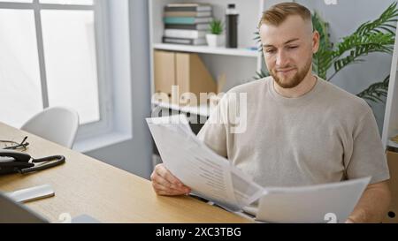 Un giovane uomo caucasico e barbuto esamina i documenti in un ambiente di ufficio moderno, evocando attenzione e professionalità. Foto Stock