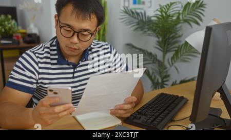 Un giovane e affascinante cinese lavora in un ufficio, controllando attentamente il suo smartphone e i documenti alla sua scrivania, indossando una camicia a righe con una com Foto Stock