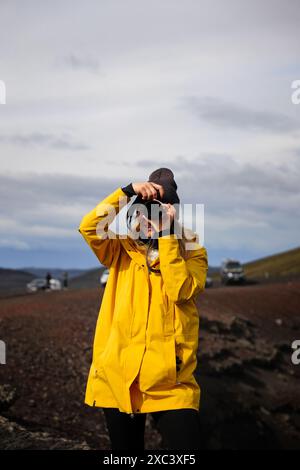 Una ragazza con zaino in spalla che scatta una foto dei suoi dintorni negli altipiani islandesi. Indossa una giacca da trekking gialla e guarda la macchina fotografica mentre h Foto Stock