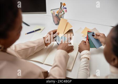 Primo piano di mani femminili che tengono in mano campioni di carta in colori pastello contro la scrivania con penne e notebook, concetto di lavoro creativo Foto Stock