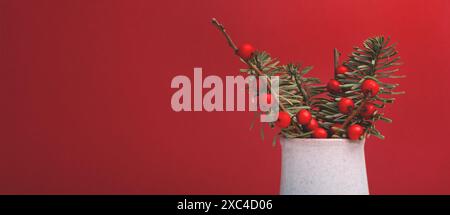 Striscione con bouquet di nobilis abete e rami di winterberry in un vaso di fronte allo sfondo rosso. Foto Stock