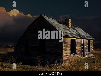 Vecchia casa che è stata abbandonata bagnata e che sta marcigendo via con una luna piena che sorge dalle nuvole dietro Juast dopo il tramonto del sole. Foto Stock