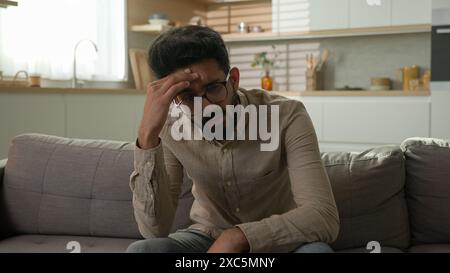 Un uomo arabo indiano penoso annoiato sul divano di casa pensando in profondità nei pensieri decisione difficile stanco stanco sofferente preoccupato sesso maschile noioso fine settimana Foto Stock