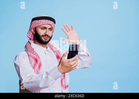 Uomo musulmano in stile tradizionale e foulard che parla in video chat su smartphone e saluta in studio. Persona araba che saluta e chatta in videoconferenza sul telefono cellulare Foto Stock