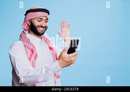 Un uomo musulmano sorridente che indossa abiti tradizionali saluta mentre parla in video chat sullo smartphone. Arabo che saluta mentre parla in una riunione virtuale sul telefono cellulare con un'espressione allegra Foto Stock