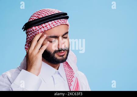 Uomo musulmano esausto con gli occhi chiusi che tiene il tempio mentre soffre di emicrania. Persona araba che indossa abiti tradizionali sentendo dolore e toccando la testa mentre ha la pressione alta Foto Stock