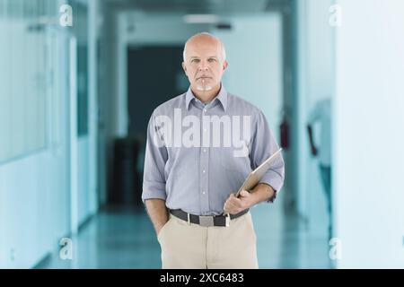 uomo calvo con un pizzetto bianco, con una camicia azzurra e pantaloni beige, si trova in un corridoio con una piegatrice. La sua espressione seria è incorniciata da TH Foto Stock