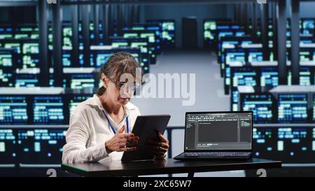 Scienziato informatico senior in server farm che completa le attività su tablet mentre esegue script su terminale notebook. Esperto tra supercomputer che manipolano il codice e migliorano la sicurezza informatica Foto Stock