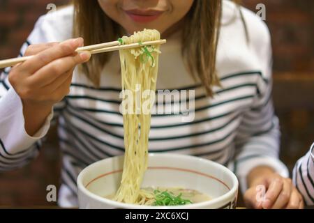 Primo piano di una donna che mangia ramen con le bacchette. La donna, indossando una camicia a righe bianca e nera, solleva una porzione di spaghetti da una ciotola Foto Stock