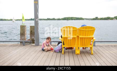 Sedie gialle Adirondack sul molo di legno che si affaccia sul lago con barche a vela e giochi per bambini - TORONTO, ON Canada - 03.29. 2024. Foto Stock