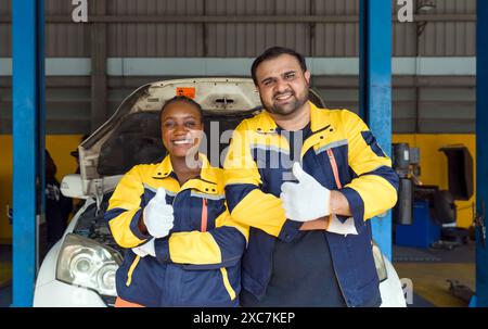 Due meccanici in uniforme da lavoro e un guanto di protezione sollevano i pollici e si alzano davanti a un'auto con cofano aperto. Foto Stock