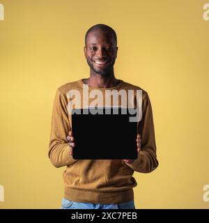 Sorridente giovane afro-americano che tiene in mano un tablet, presenta il dispositivo su uno sfondo giallo - uomo felice che mostra lo schermo digitale - moderno Technolo Foto Stock