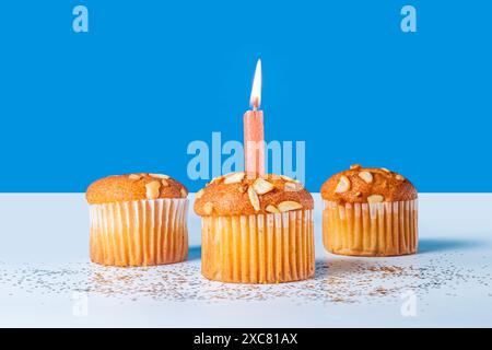 Tre cupcake di compleanno con una candela illuminata su un tavolo bianco su sfondo blu Foto Stock