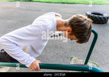 Un giovane con una camicia bianca sta facendo un esercizio su una serie di barre parallele in una palestra all'aperto. Sta usando le braccia per spingersi su e giù, Foto Stock