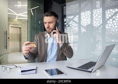 Uomo d'affari preoccupato in un ufficio moderno, con una carta di credito mentre parla al telefono e guarda il suo portatile con preoccupazione. Foto Stock