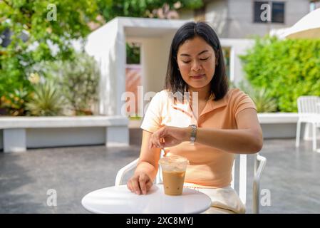 Donna seduta in una caffetteria all'aperto. È vestita in modo informale con un top arancione e si gode un caffè ghiacciato posto sul tavolo di fronte a lei. La S Foto Stock