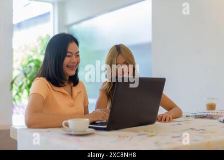 Due donne sedute al bar che usano un computer portatile insieme mentre sorridono felicemente Foto Stock