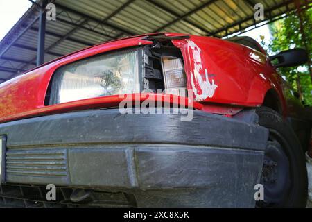 Vista dal basso verso l'alto della parte anteriore di un'auto con un faro danneggiato e graffi - primo piano. Una vecchia auto rossa brillante con danni. Foto Stock