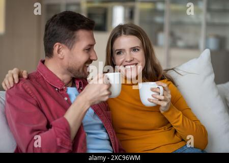 Amata coppia uomo donna che passa un fine settimana pigro beve un caffè seduto sul divano a casa, divertitevi a parlare. Foto Stock