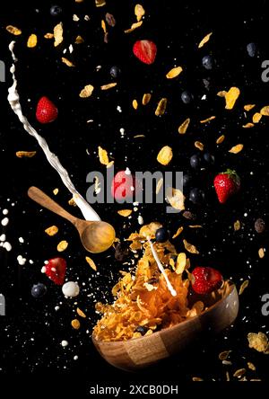 I fiocchi di mais, il latte, le fragole e i mirtilli volano fuori da una ciotola su uno sfondo nero Foto Stock