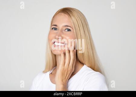 Donna bionda sorridente che tocca il suo volto su Uno sfondo bianco Foto Stock