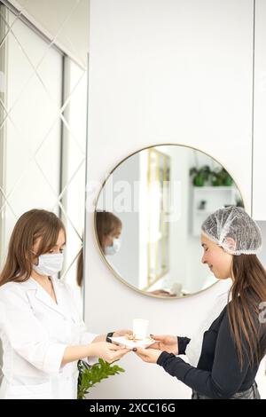 Il dermatologo che indossa una tuta bianca e una maschera facciale sta servendo un cliente con una tazza di caffè e alcuni dolci su un piatto da portata. Il cliente lo è Foto Stock