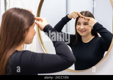 Una giovane e bella donna nell'ufficio del dermatologo si guarda allo specchio e sorride mentre stringe un brufolo sulla fronte. Foto Stock