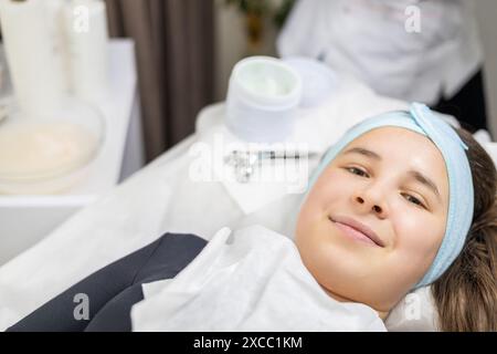 Primo piano sorridente giovane donna in attesa di una procedura di bellezza, sdraiata su un tavolo di trattamento, vicino a una maschera facciale e un rullo di massaggio facciale. Foto Stock