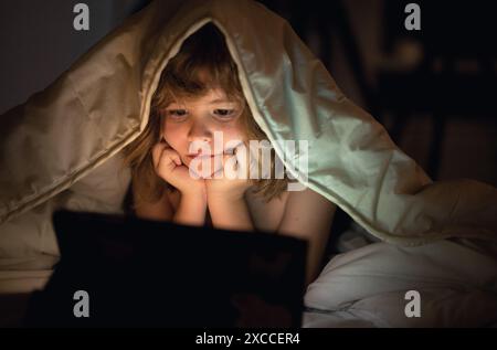 Bambino che guarda il tablet prima di dormire. Il bambino legge l'e-book. Il ragazzino guarda lo schermo del tablet in alto. Ragazzo che gioca un tablet sdraiato su un Foto Stock