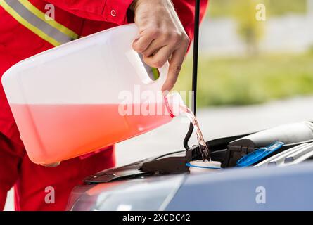 Operatore addetto alla manutenzione da vicino, conducente che versa liquido lavavetri per la pulizia del parabrezza antigelo nel serbatoio dell'auto del cofano dalla bottiglia del filtro a carboni attivi. Detergente liquido per vetri per parabrezza per uso estivo o invernale Foto Stock