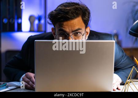 Un uomo d'affari indiano fiducioso che si nasconde dietro un computer portatile, guarda la fotocamera, spiare i colleghi che lavorano, sbirciare. Un freelance professionista che guarda da dietro il computer, sguardo astuto Foto Stock