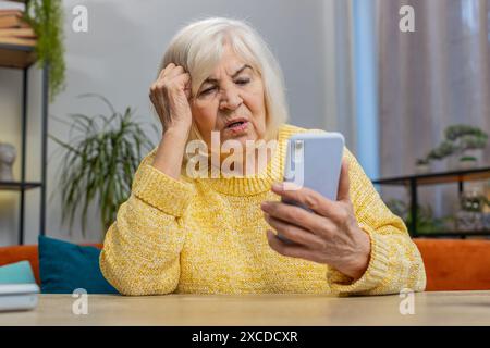 Una donna anziana triste usa la digitazione dello smartphone sfogliando, perde diventando sorpreso risultati improvvisi della lotteria, cattive notizie, perdita di fortuna, fallimento. La nonna anziana nel salotto di casa si siede sul divano alla scrivania Foto Stock