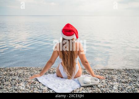 Una donna che indossa un cappello di Babbo Natale rosso si siede su un telo da spiaggia. Indossa un bikini e ha un portatile in grembo Foto Stock