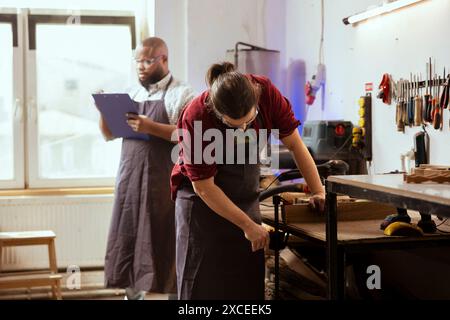 Lavoratore di legno che indossa occhiali di sicurezza mentre utilizza una morsa da banco per tenere il blocco di legname in officina accanto al collaboratore. Artigiano che utilizza dispositivi di protezione e morsa per bloccare il pezzo di legno Foto Stock