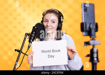 Donna allegra che tiene in mano un cartello con il messaggio World Book Day, filma con cellulare, sfondo studio. Creatore di contenuti allegri che registra video con lo smartphone, promuovendo l'importanza della lettura Foto Stock