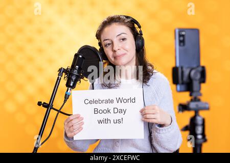 Persona allegra che tiene in mano un cartello con il messaggio del giorno del libro mondiale, filma con telefono e microfono, sfondo studio. Video di registrazione radiosa di librai che promuovono l'importanza dell'alfabetizzazione Foto Stock