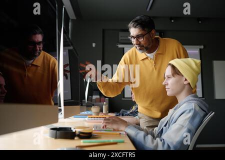 Insegnante birazziale maturo di informatica che spiega il soggetto a uno dei suoi studenti durante la lezione in classe moderna Foto Stock