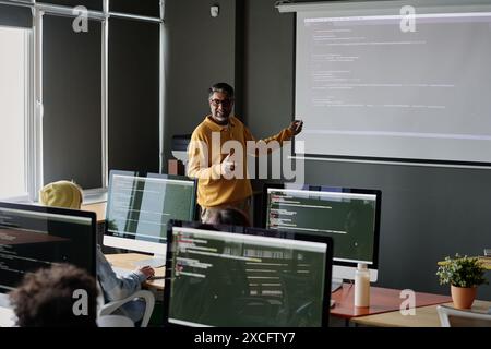 Allegro e maturo insegnante di sesso maschile in piedi di fronte agli studenti che spiega i problemi di codifica durante la lezione di informatica Foto Stock