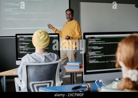 Uomo birazziale maturo che indossa abiti casual intelligenti che insegnano a programmare in una moderna classe dotata di computer Foto Stock