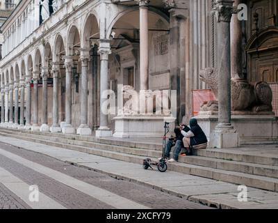 Cremona, Italia - 10 giugno 2024 due persone fanno una pausa sui gradini di un edificio storico con uno scooter elettrico parcheggiato accanto a loro Foto Stock