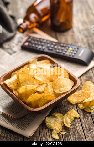 Una ciotola piena di patatine si trova su un tavolo di legno, accanto a un telecomando della televisione e a due bottiglie di birra. L'attenzione è sulla ciotola di patatine, che a Foto Stock